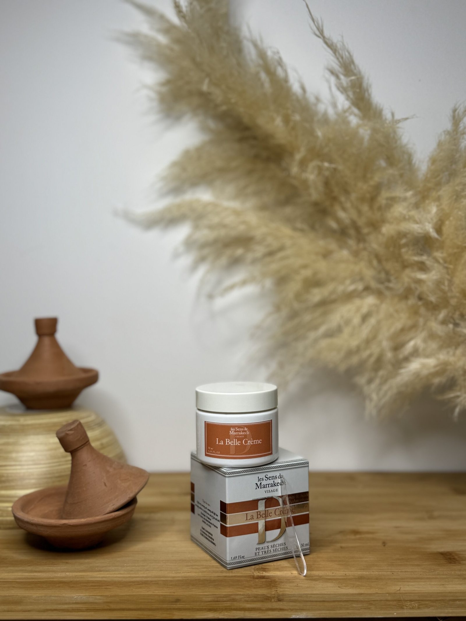 Pot et boîte de La Belle Crème Les Sens de Marrakech posés sur une surface en bois, avec tajines en terre cuite et herbes de pampa en arrière-plan