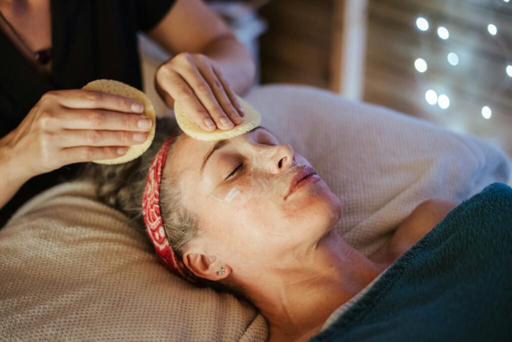 Adult lady lying in spa center while massager removing mask from face with sponge
